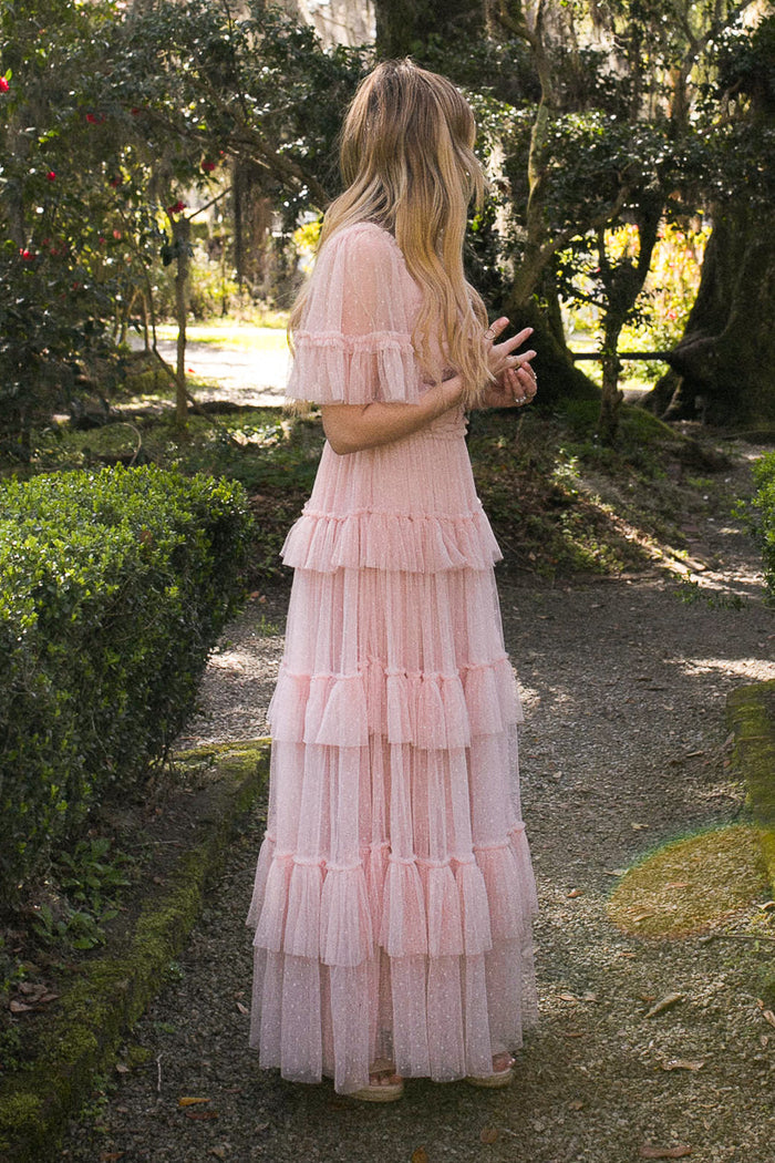 A woman with long, wavy blonde hair stands sideways on a garden path surrounded by greenery, wearing the Ivy City Co Whimsical Dress in Blush—a floor-length, ruffled blush pink dress with short sleeves.
