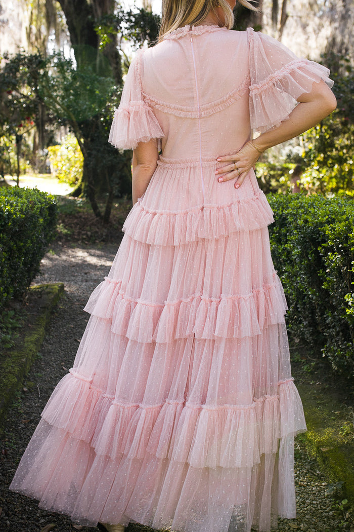 A woman stands on a garden path surrounded by greenery, her back to the camera as she wears Ivy City Co’s Whimsical Dress in Blush, featuring tiered ruffles, short sleeves, and a fitted waist.