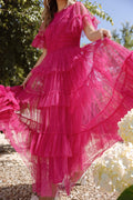 A woman stands outdoors on a sunny day, surrounded by greenery and white flowers, showing off the volume of her Ivy City Co Whimsical Dress in Fuchsia with ruffled, sheer tulle skirt and layered tiers. Her face is not visible.