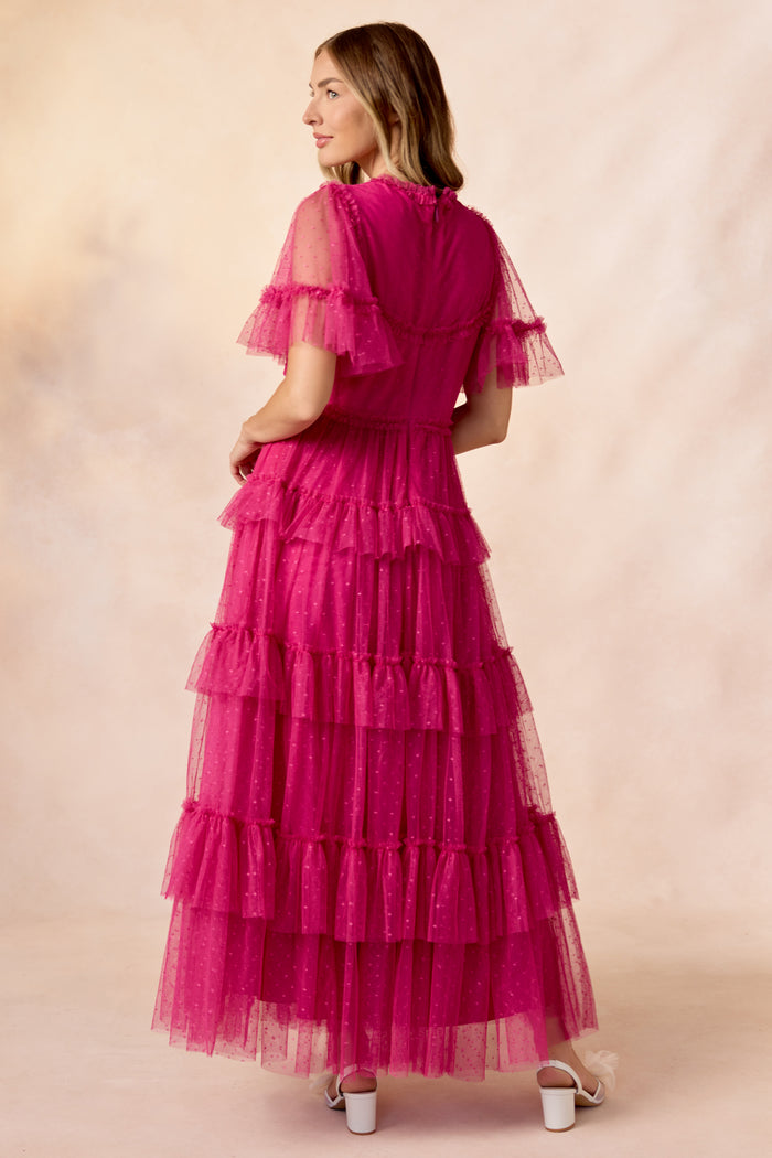 A woman with shoulder-length hair and white heeled sandals faces away, wearing the Ivy City Co Whimsical Dress in Fuchsia—featuring tiered tulle, ruffled sleeves, and polka dot details—set against a soft, light backdrop.