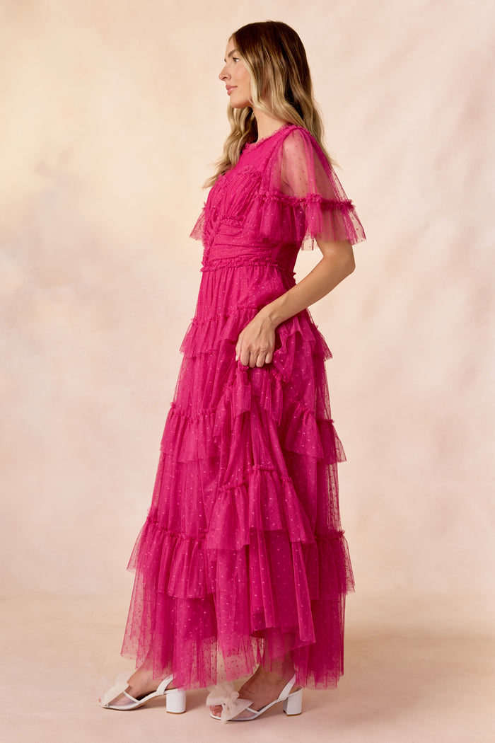 A woman in profile wears the Whimsical Dress in Fuchsia by Ivy City Co, featuring a tiered tulle skirt and sheer short sleeves, paired with white heels against a soft beige and pink backdrop.