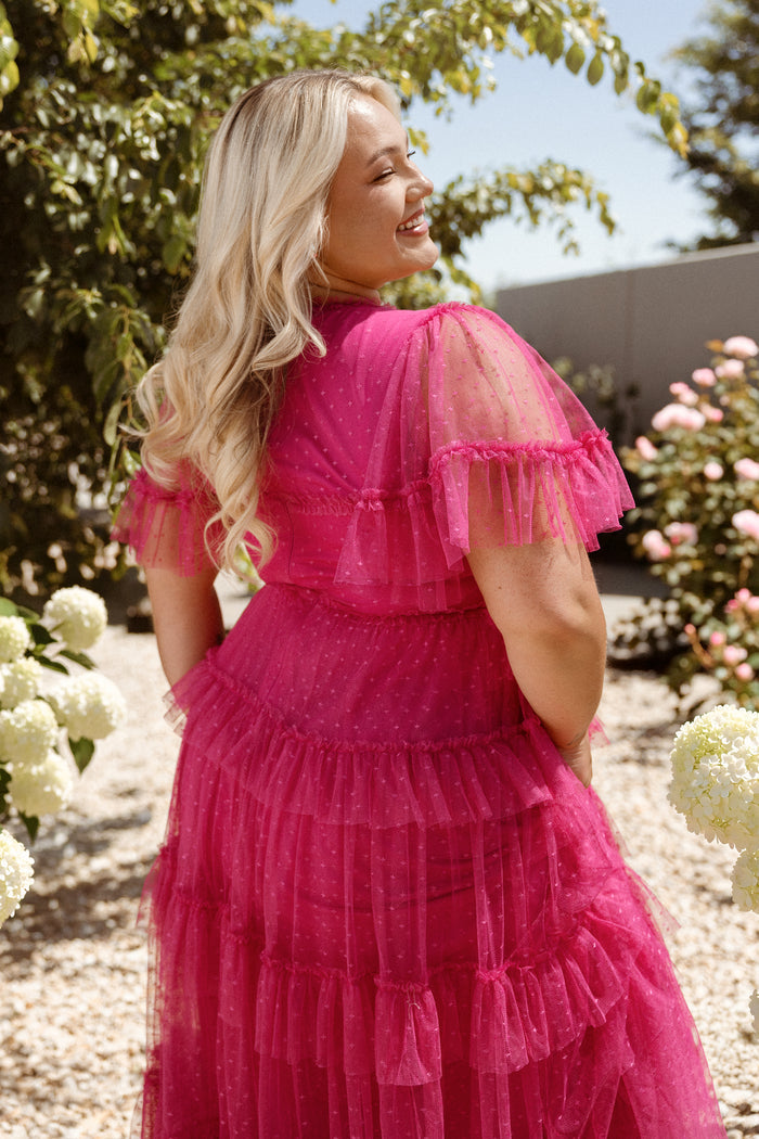 A woman with long blonde hair, smiling over her shoulder, stands in a sunny garden surrounded by white and pink flowers, wearing the Whimsical Dress in Fuchsia by Ivy City Co—a bright dress with a ruffled tulle skirt.