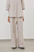 A person wearing a light beige matching two-piece outfit with a short-sleeve button-front top and wide-leg pants, standing against a plain light gray wall and floor.